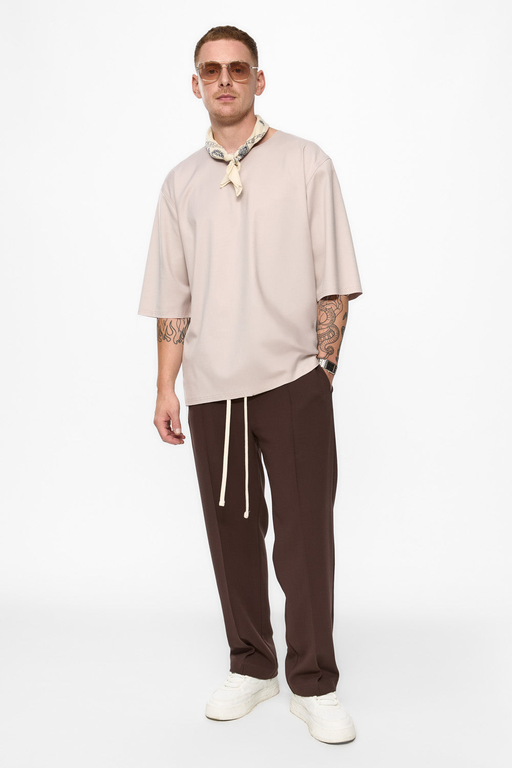 Man wearing a beige oversized shirt, brown pants, and white shoes on a white background