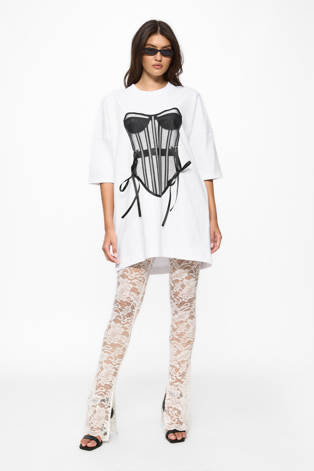 Woman wearing a white oversized shirt with a black corset graphic and lace pants on a white background